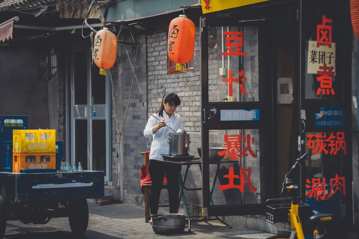 A female cook preparing food outdoors in a Beijing hutong, with traditional decor and lanterns.