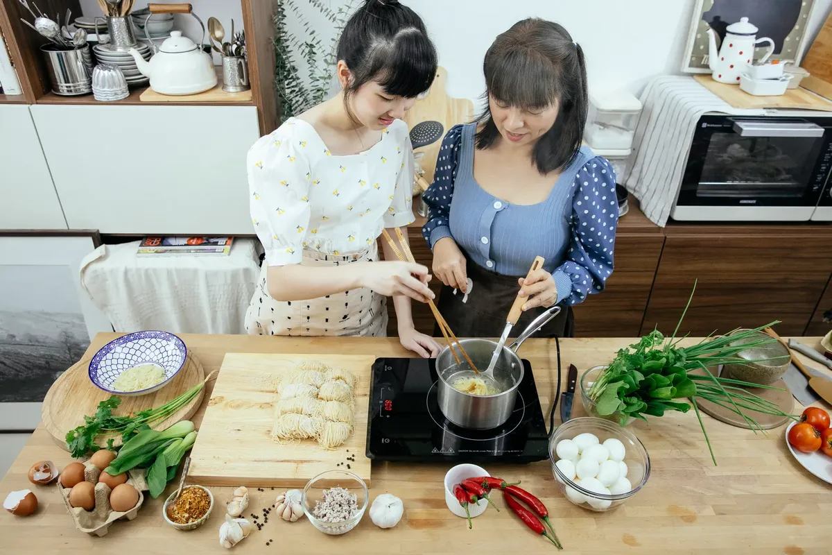 High angle glad Asian females in stylish aprons with chopsticks boiling homemade noodles in saucepan on tabletop stove while cooking Asian dish together in cont